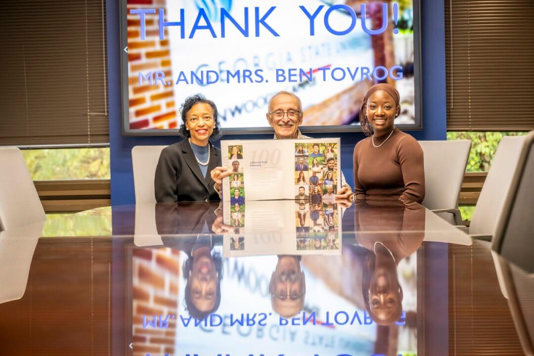 Two women and one man sit at a table holding up a book.