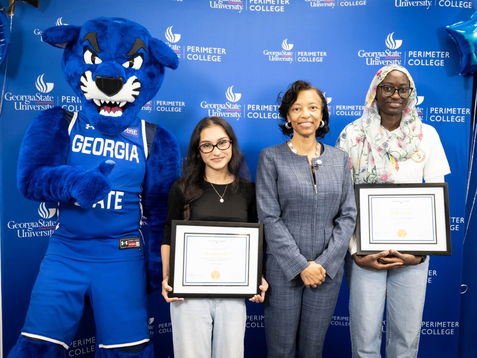 Pounce (Georgia State University mascot - blue panther) stands with Dean of College and two students holding framed certificates.