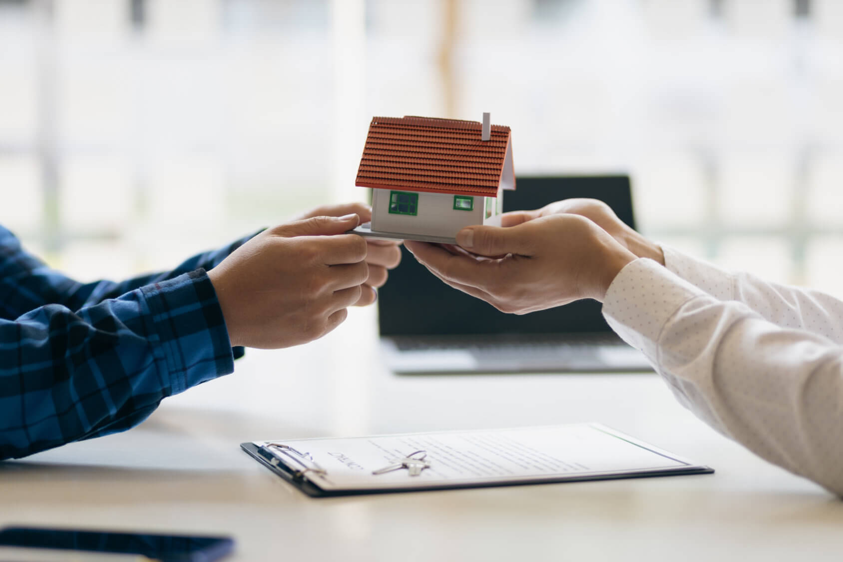 Image of a small house being held up by two sets of hands on either side. Underneath them is a clipboard with paperwork and a key on top.