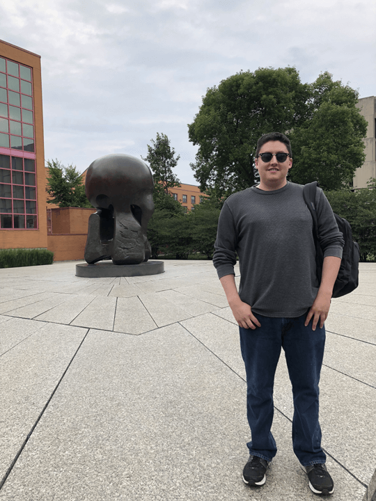 Marc Reyes standing with sunglasses and a backback in front of a statue representing the first human-created sustained nuclear reaction.