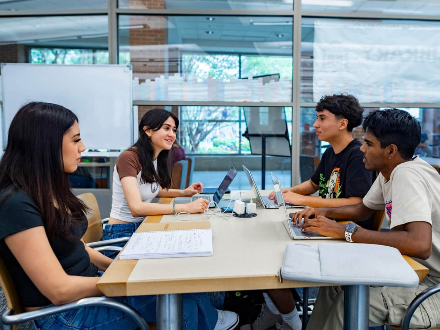 four students sit at a table with laptops studying
