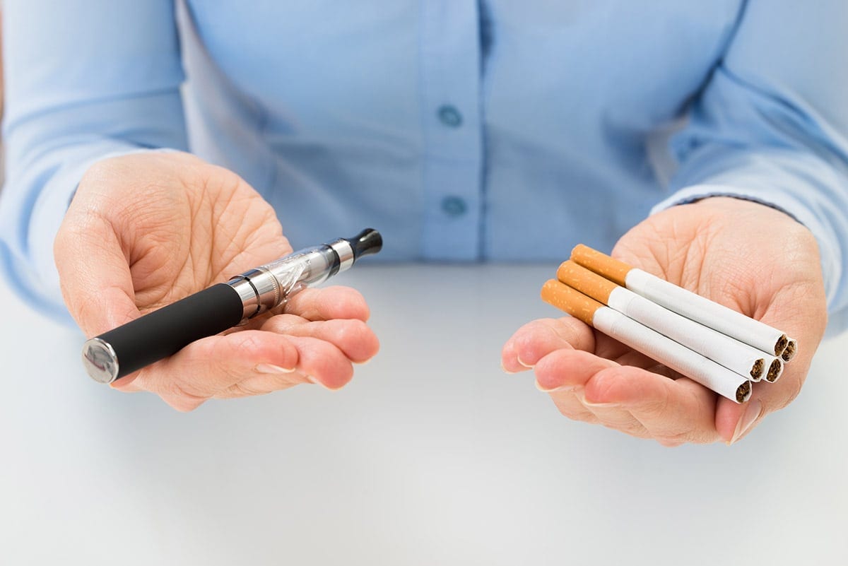 A woman holds an e-cigarette in one hand and several cigarettes in another