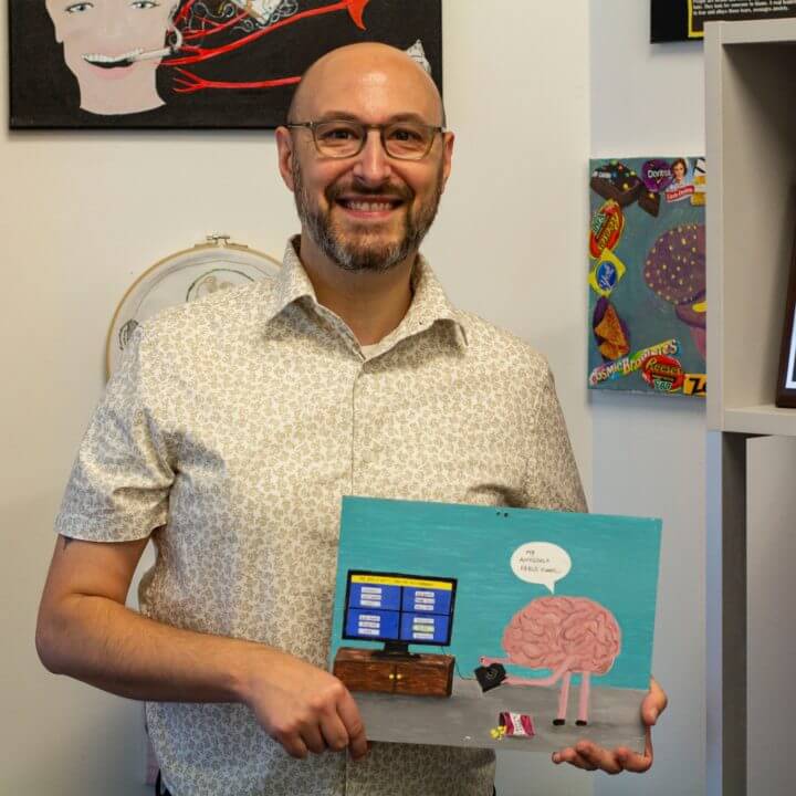 This is a photo of Dr. Joe Normandin holding a piece of artwork. The artwork is a brain talking to a computer.