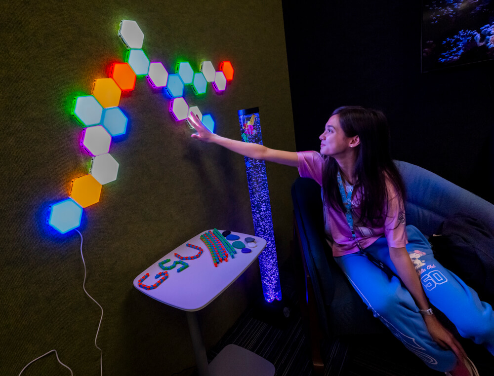 Young woman in her 20s sitting in chair touching a series of lights on the wall.