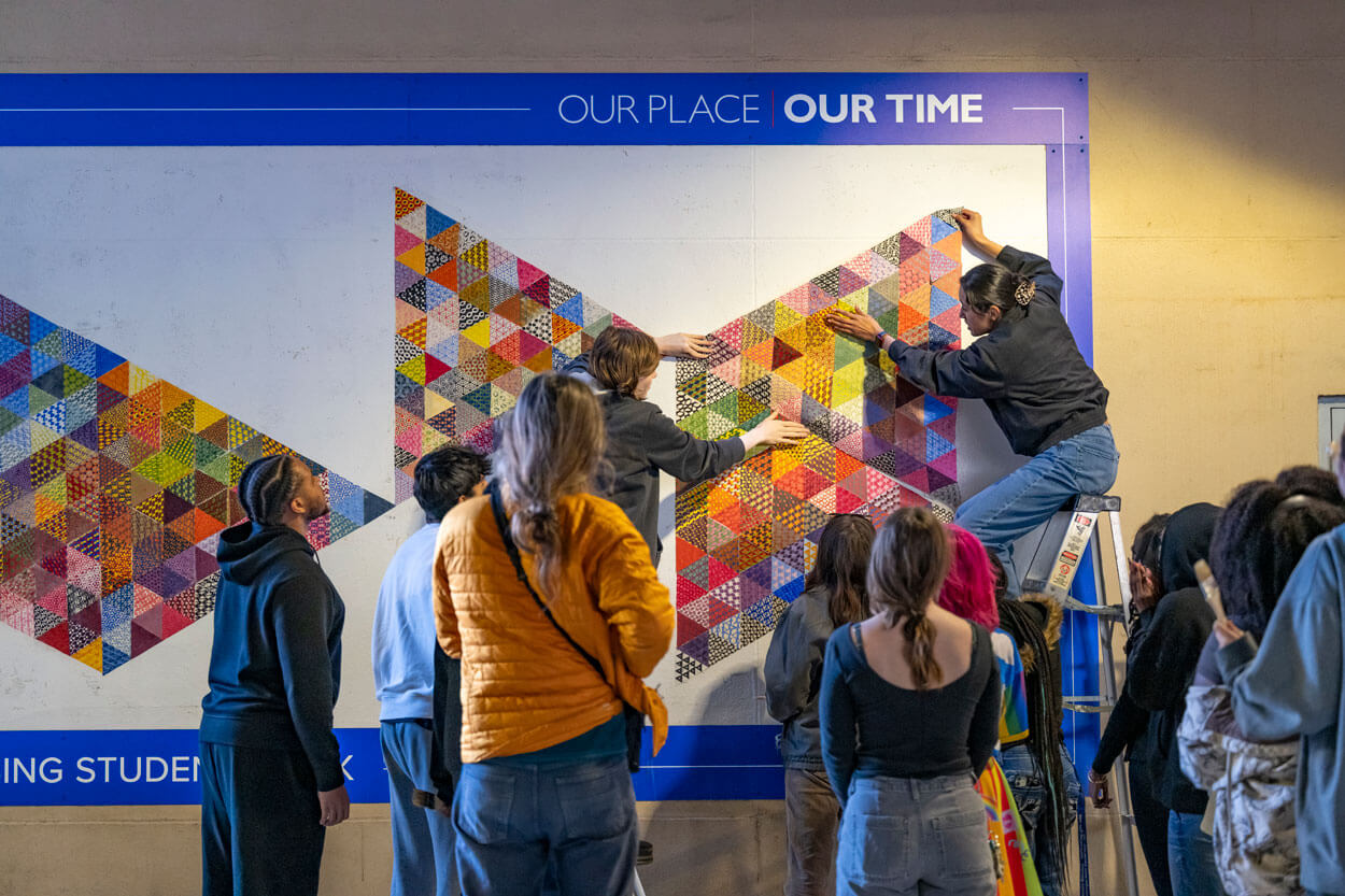 Professors watch as students install mural panels.
