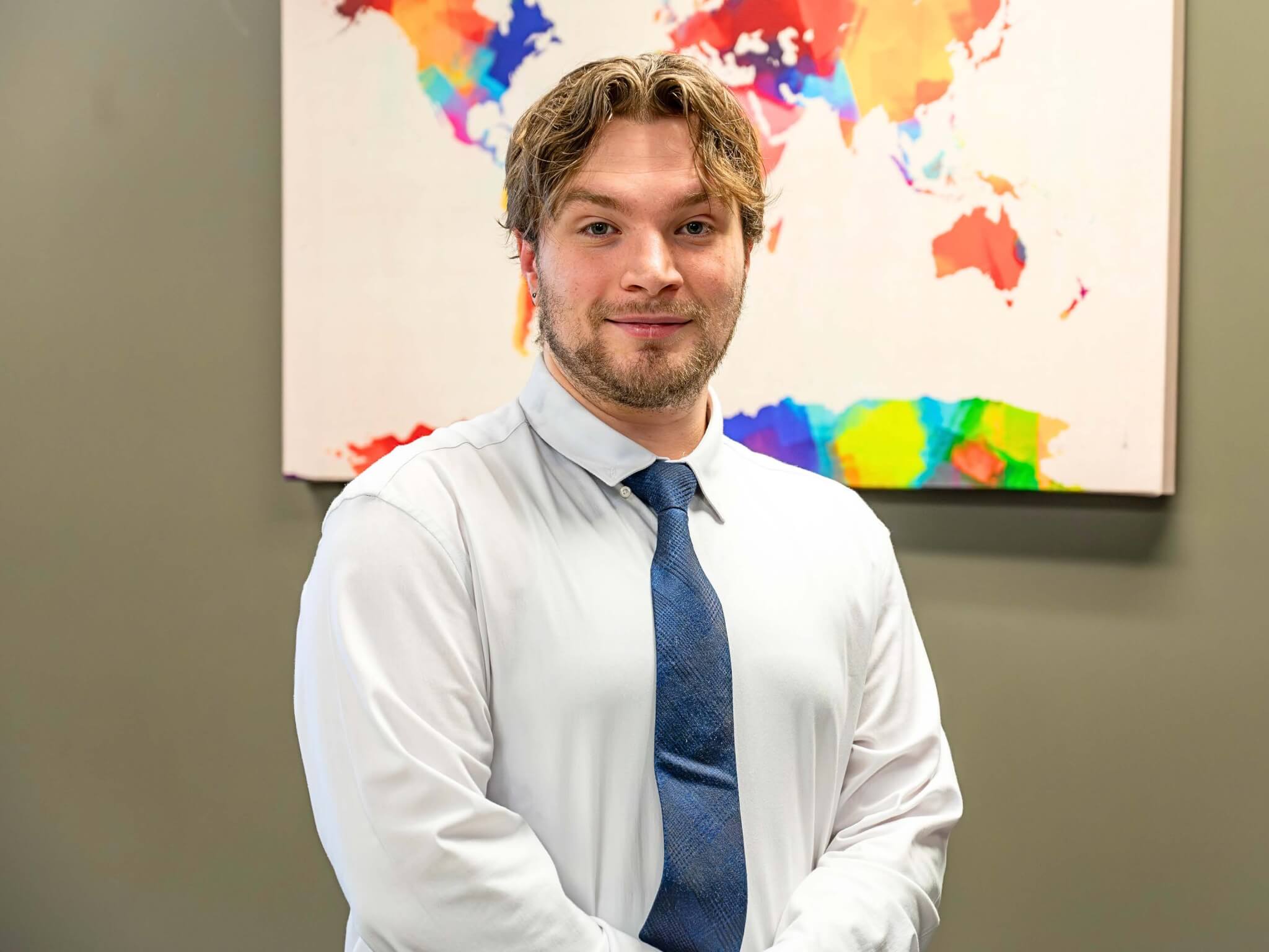 Tyler Lewchuck, a student of world languages and cultures, smiles in a white shirt and blue tie while standing in front of a colorful world map painting.