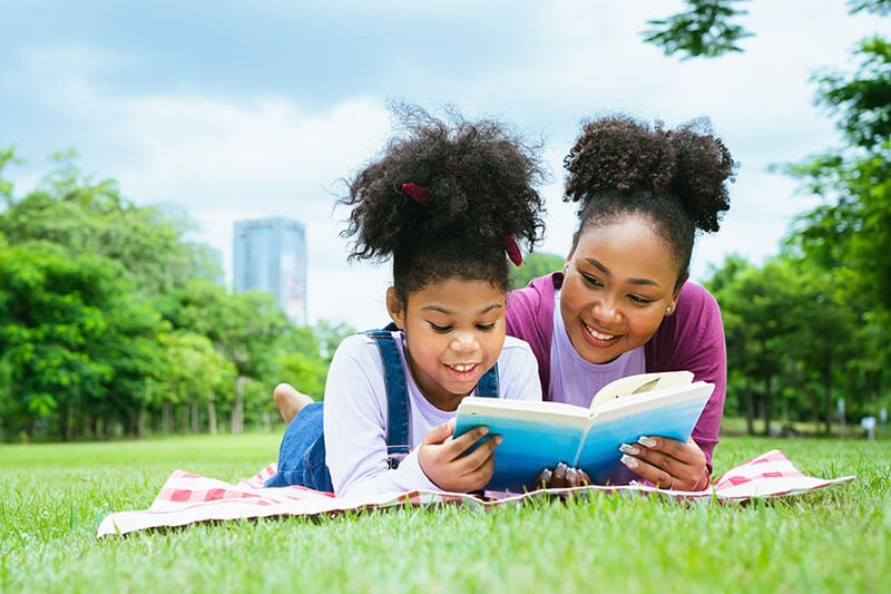 Mother and daughter lying on a blanket in a park, reading a book together on a sunny day.