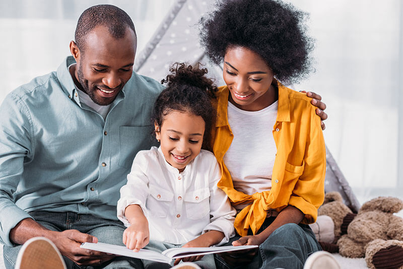 Smiling parents and their daughter reading a book together in a cozy, bright room.
