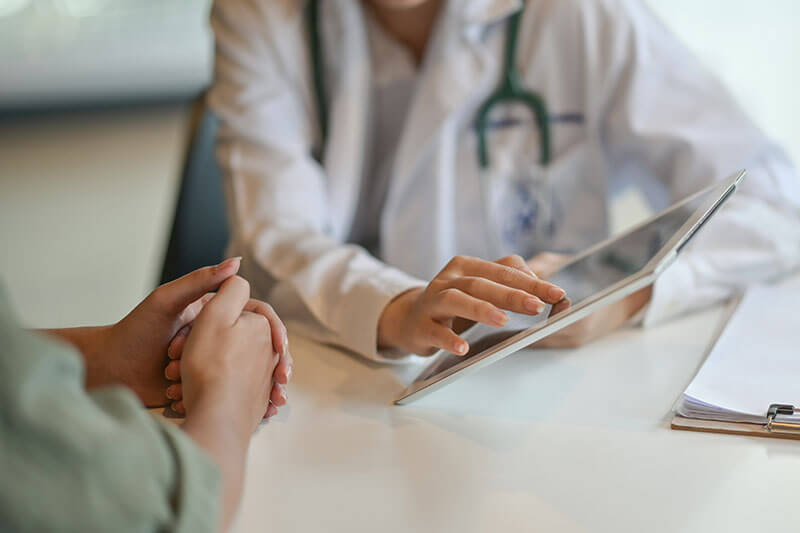 Doctor in white coat using a tablet to explain medical information to a patient sitting across the table.