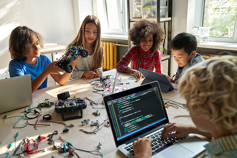 Children collaborating on a robotics project with laptops and electronic components spread across a bright workspace.